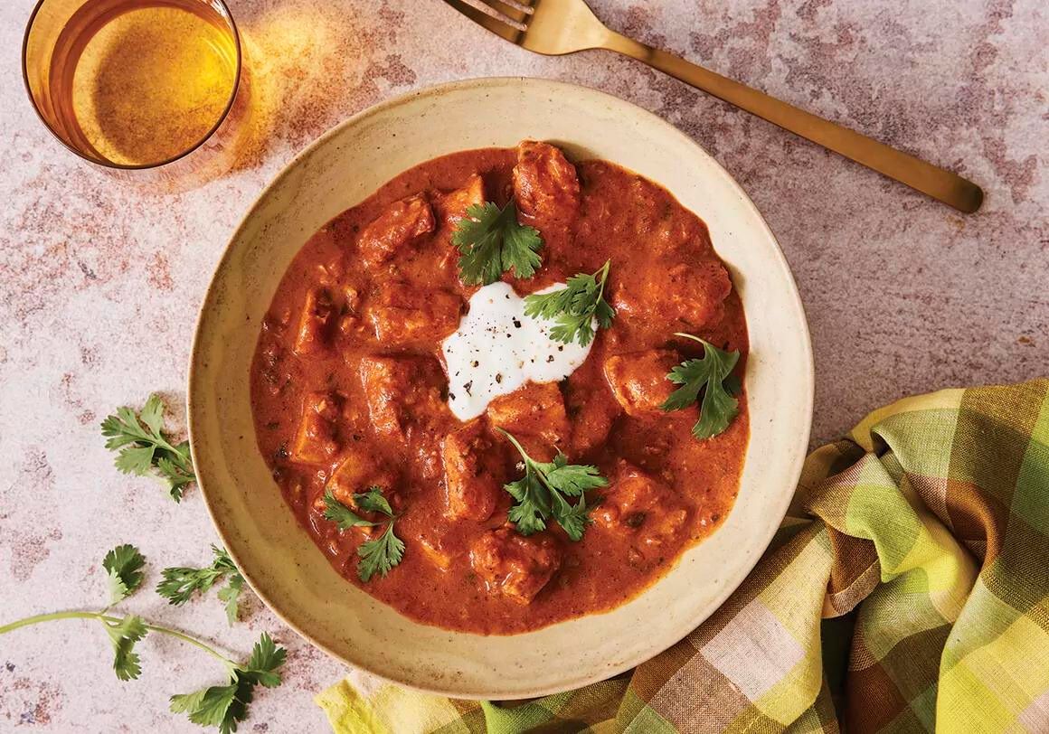 A bowl of Chicken Tikka Masala with coriander and yogurt garnish on a pink table with a glass of water, gold fork and checked napkin