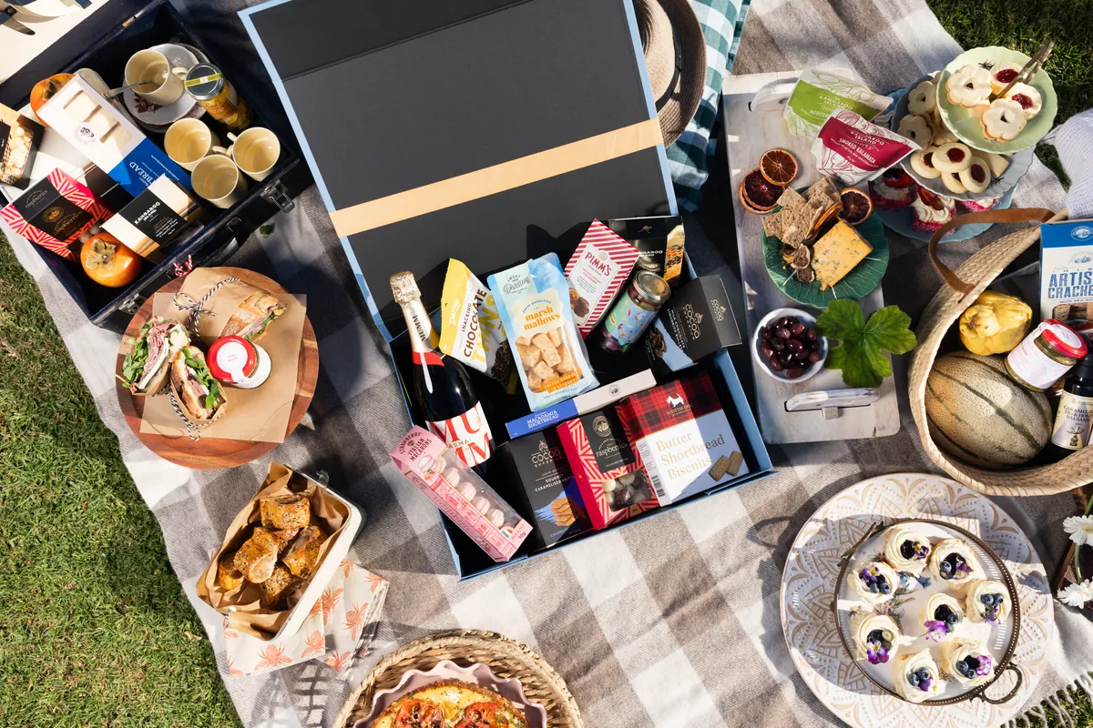 An overhead spread of a picnic hamper with Mumm sparkling wine, butter shortbread biscuits, chocolate, marshmallows and raspberries, surrounded by cheese, crackers, olives and sandwiches on a picnic rug