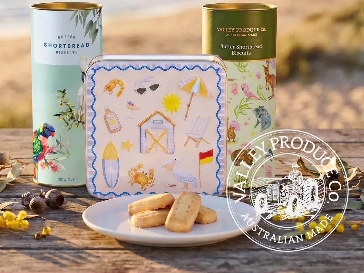 Valley Produce Co. Butter Shortbread Biscuits in a beach-themed tin and two gift tubes, with a plate of shortbread fingers, on a timber table