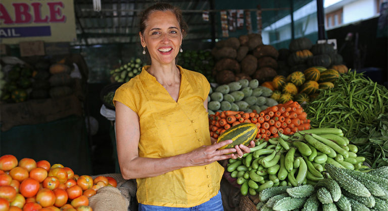 Maya Kaimal standing in a fresh food market with colourful vegetables
