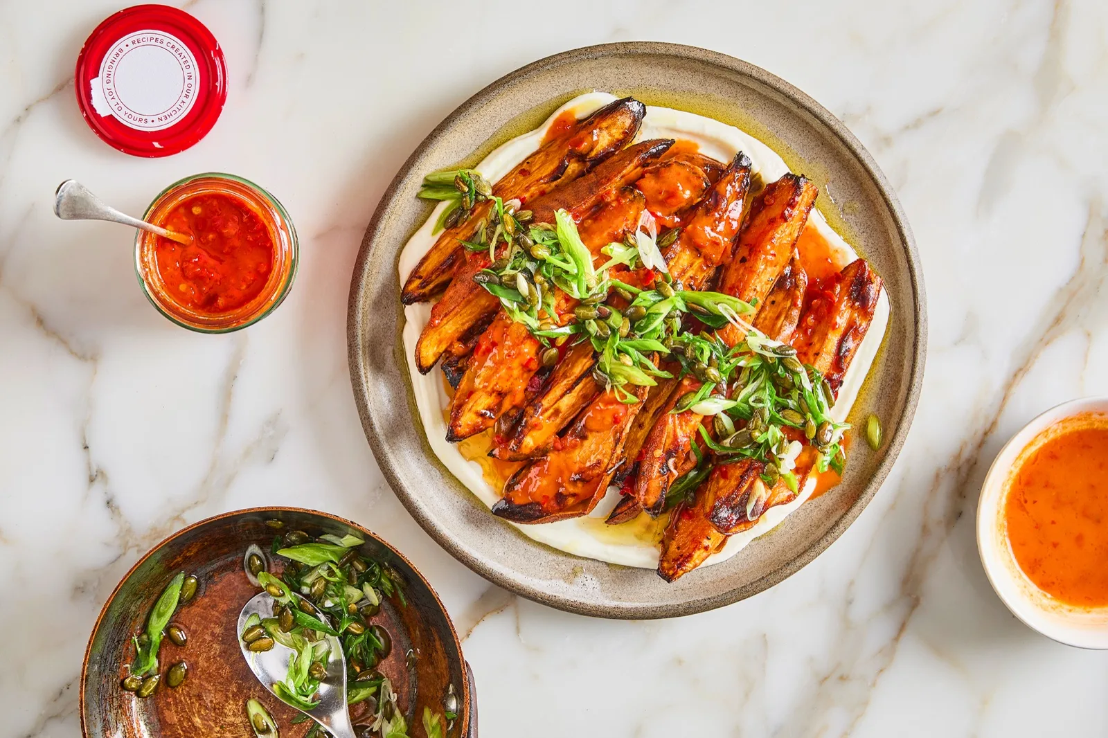 Open jar of harissa style chilli paste beside roasted sweet potato wedges with spring onion and seeds