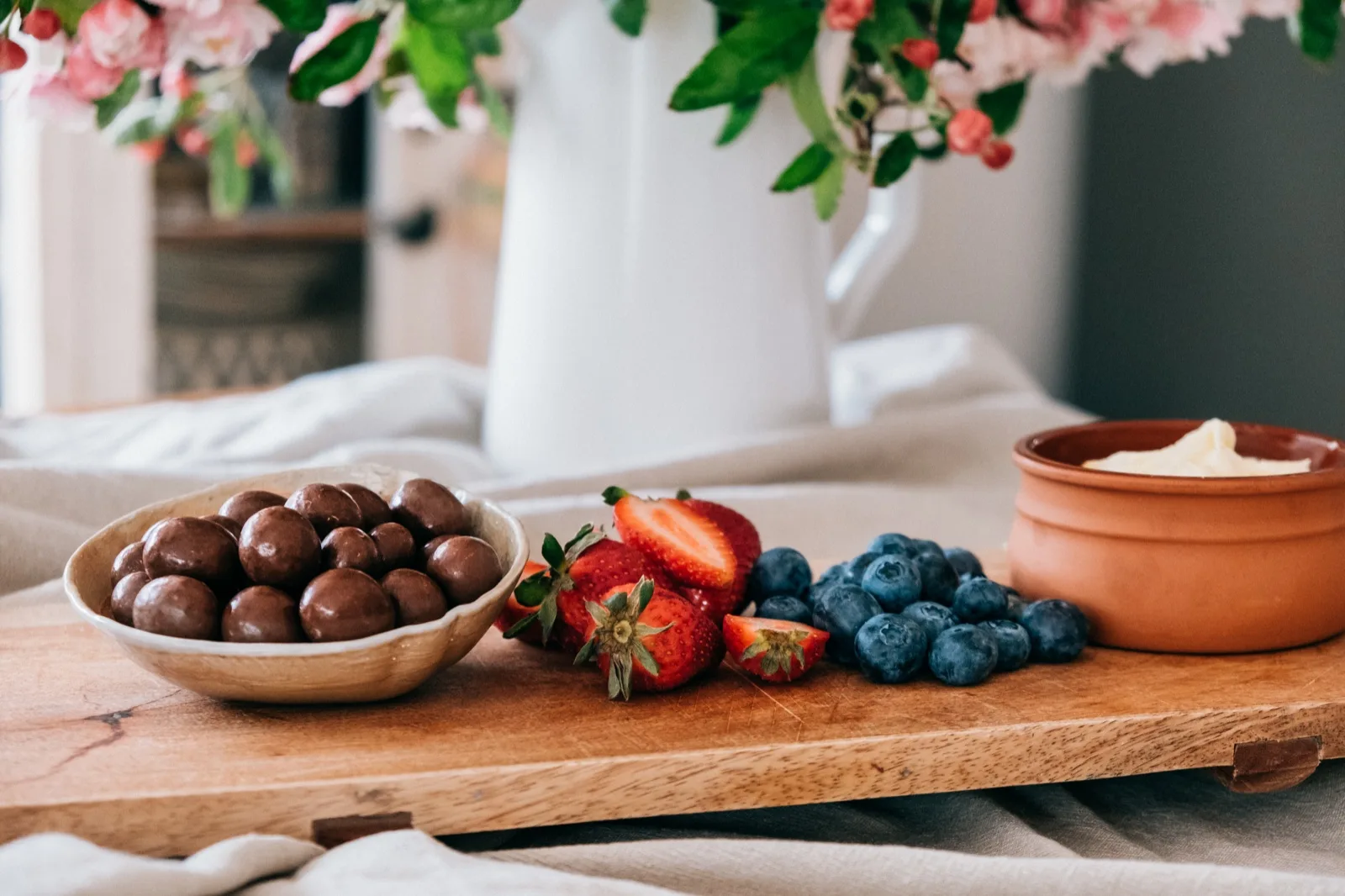 Halved strawberries, blueberries and a pot of whipped cream on a wooden board in natural l