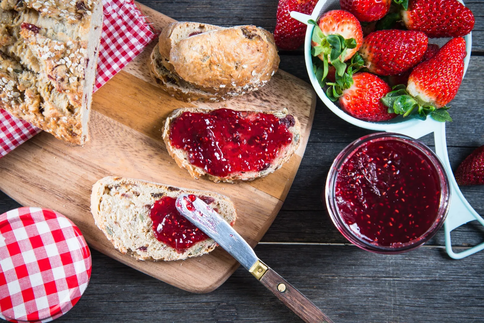 Breakfast table with sliced fruit loaf, raspberry jam spread on bread and a bowl of fresh strawberries