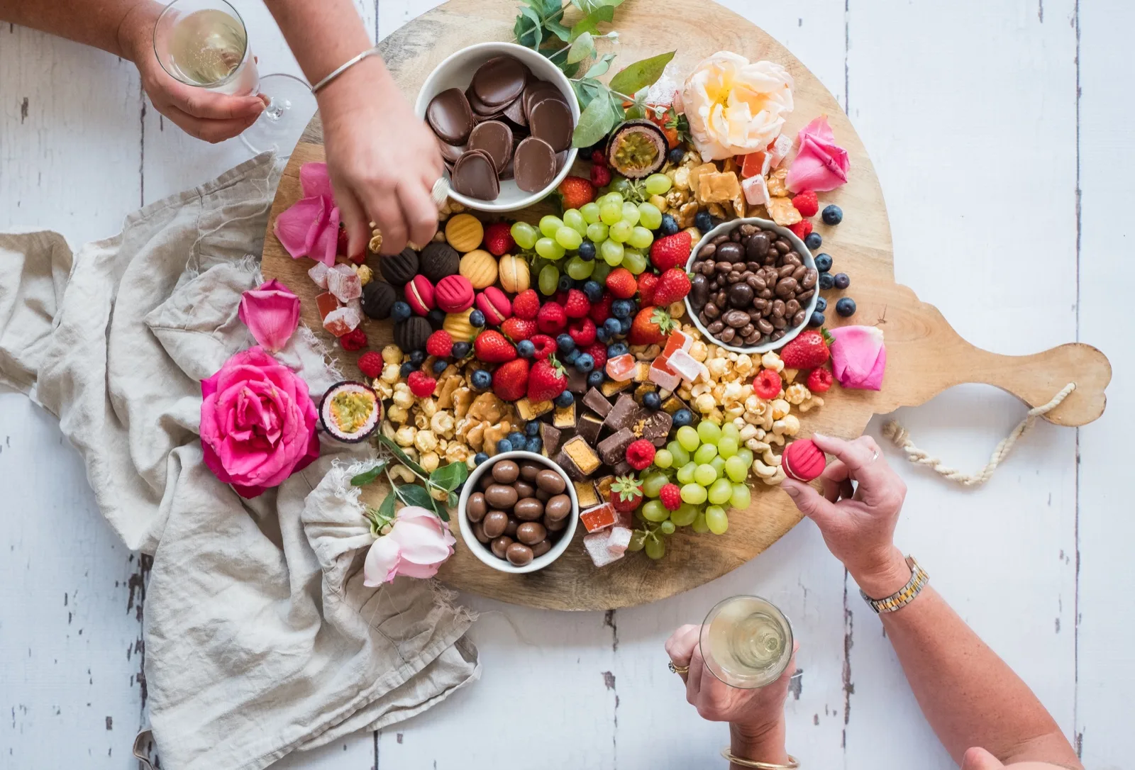 Two guests hold wine glasses and reach for macarons and chocolates on a shared board of one bite pieces