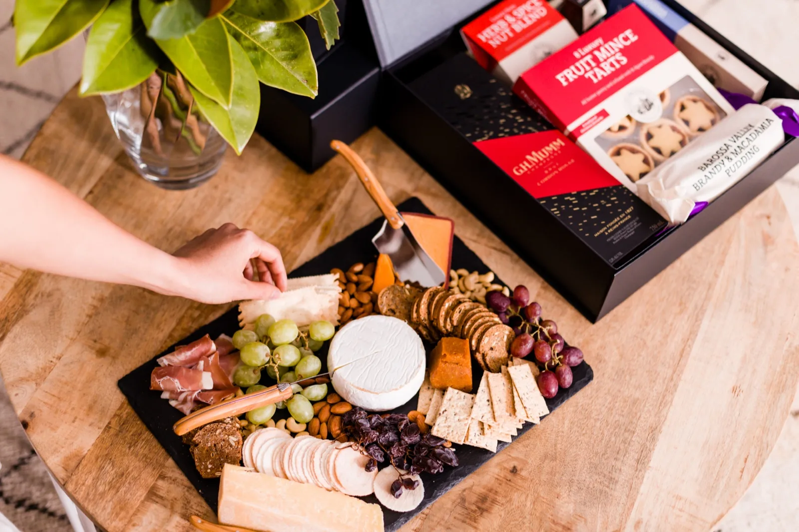 Christmas cheese board with brie, prosciutto, grapes and crackers beside an open hamper of fruit mince tarts