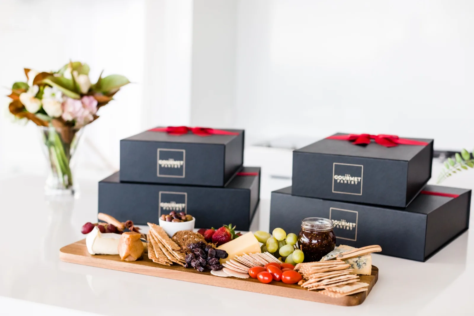 Board of crackers, brie, blue cheese, olives, quince paste and an open jar of fig jam beside sealed gift boxes
