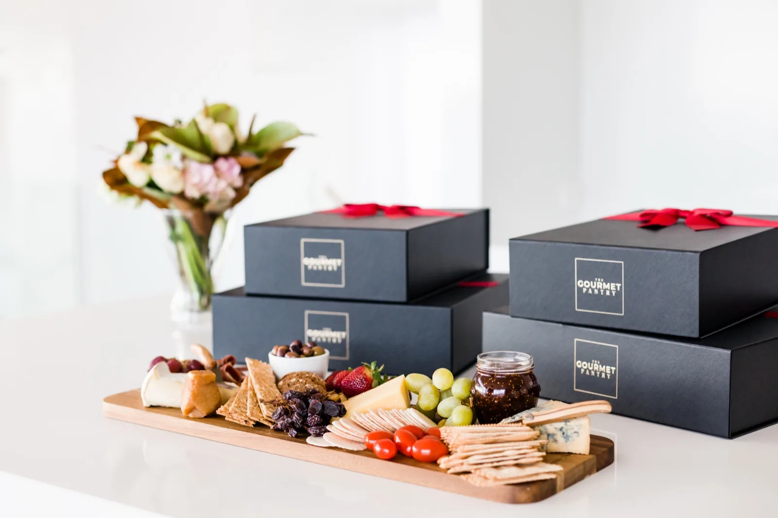 The Gourmet Pantry gift boxes stacked beside a board of cheese, crackers, olives, grapes and fig jam