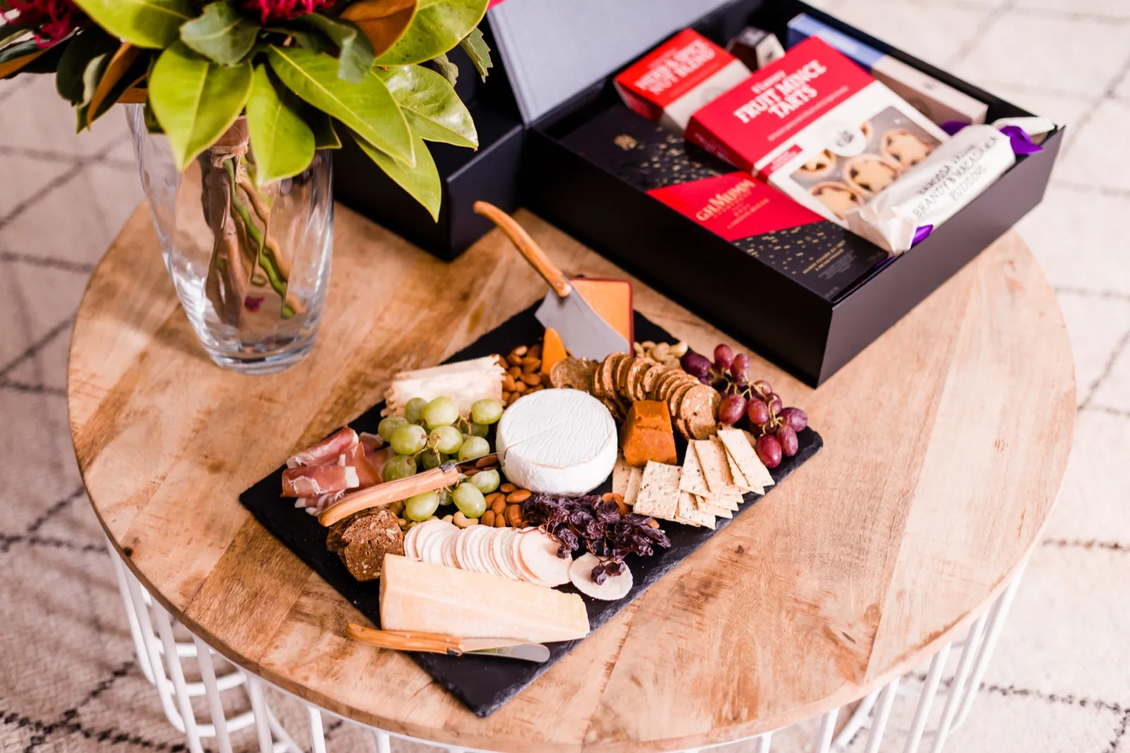 Christmas cheese board with brie, cheddar, prosciutto and grapes beside a gift box of fruit mince tarts