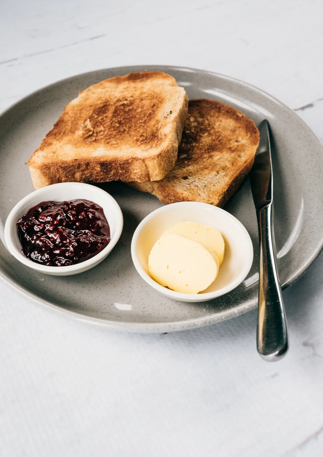 Two slices of golden toast on a grey plate beside a dish of dark red raspberry preserve an