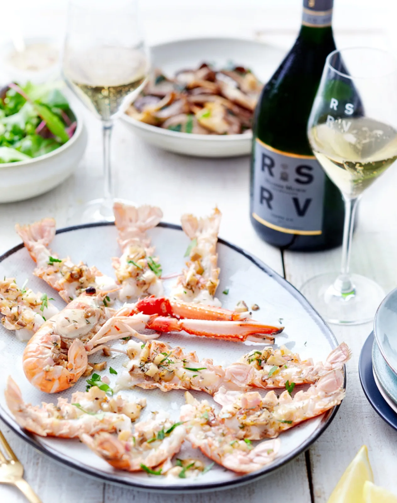 Cooked butterflied prawns topped with garlic and herbs on a platter with lemon, salad and sparkling wine