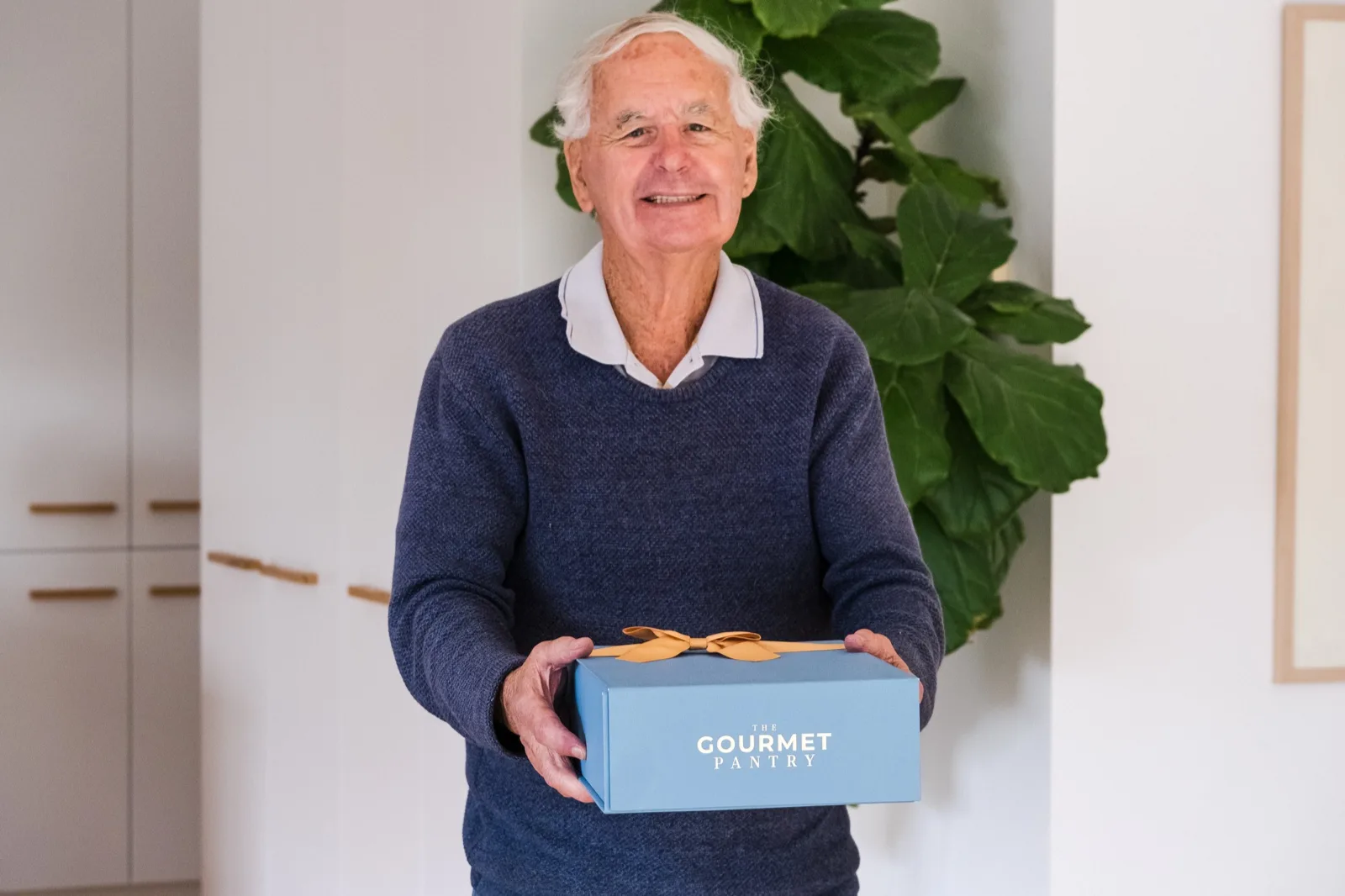 An older man in a navy jumper holding out a pale blue Gourmet Pantry gift box tied with a gold ribbon