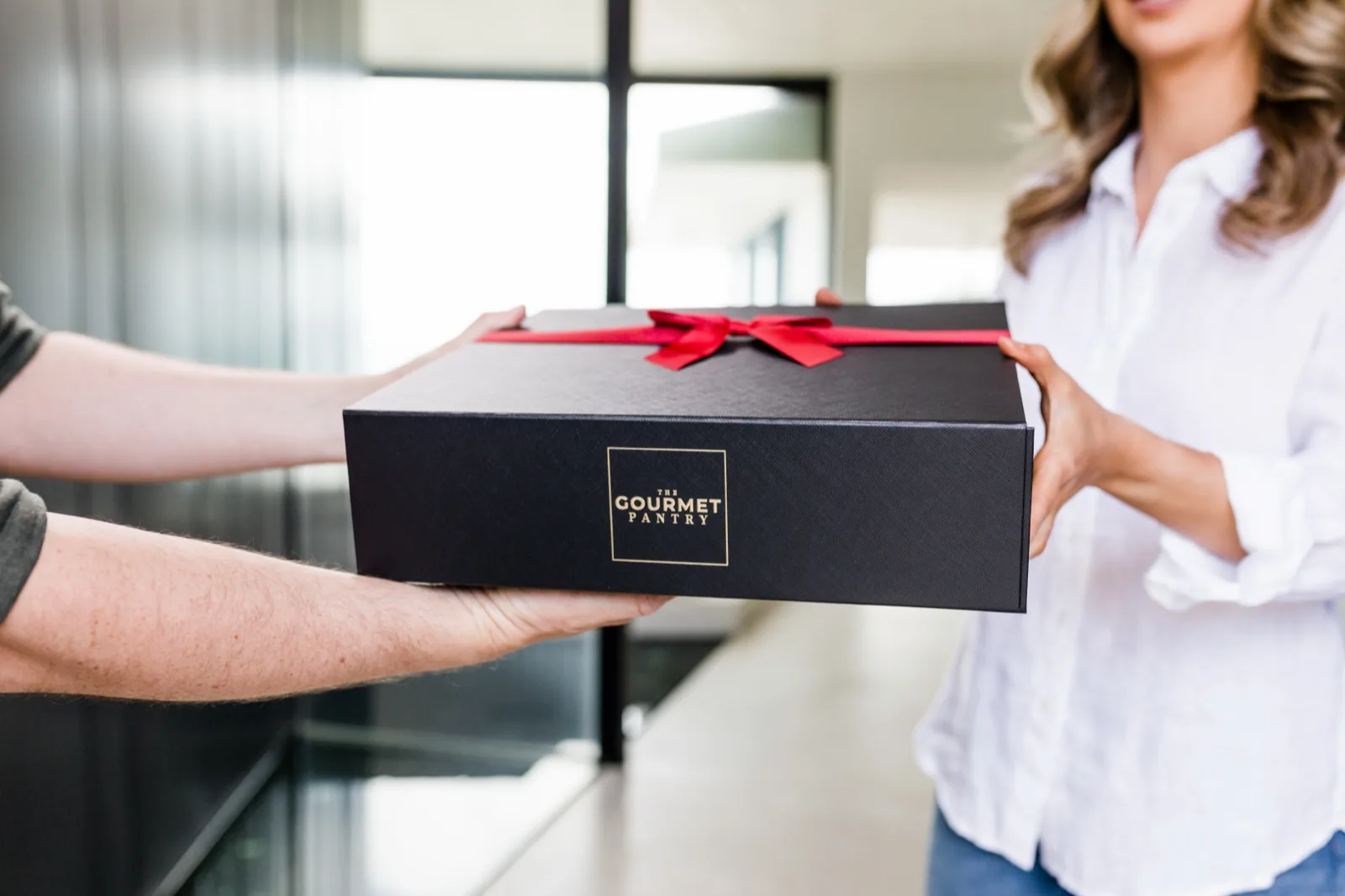 A black gift box from The Gourmet Pantry tied with a red ribbon being handed over to a woman at the door