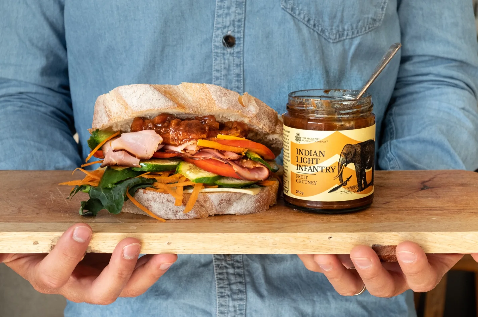 A leg ham, cheese and salad roll on a wooden board beside an open jar of fruit chutney
