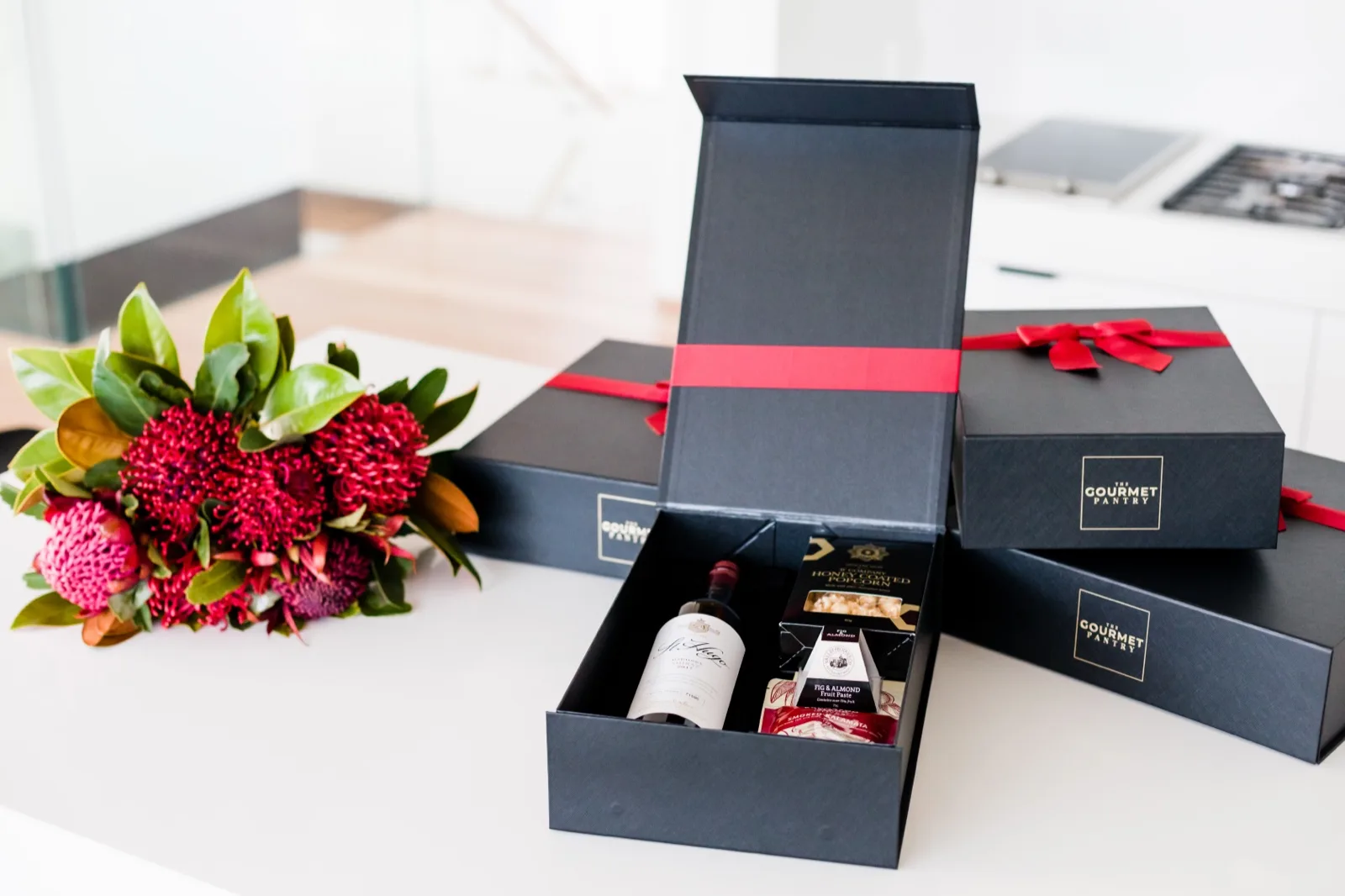 Three navy Gourmet Pantry Christmas gift boxes with red ribbons, one open showing shiraz, popcorn and fruit pa