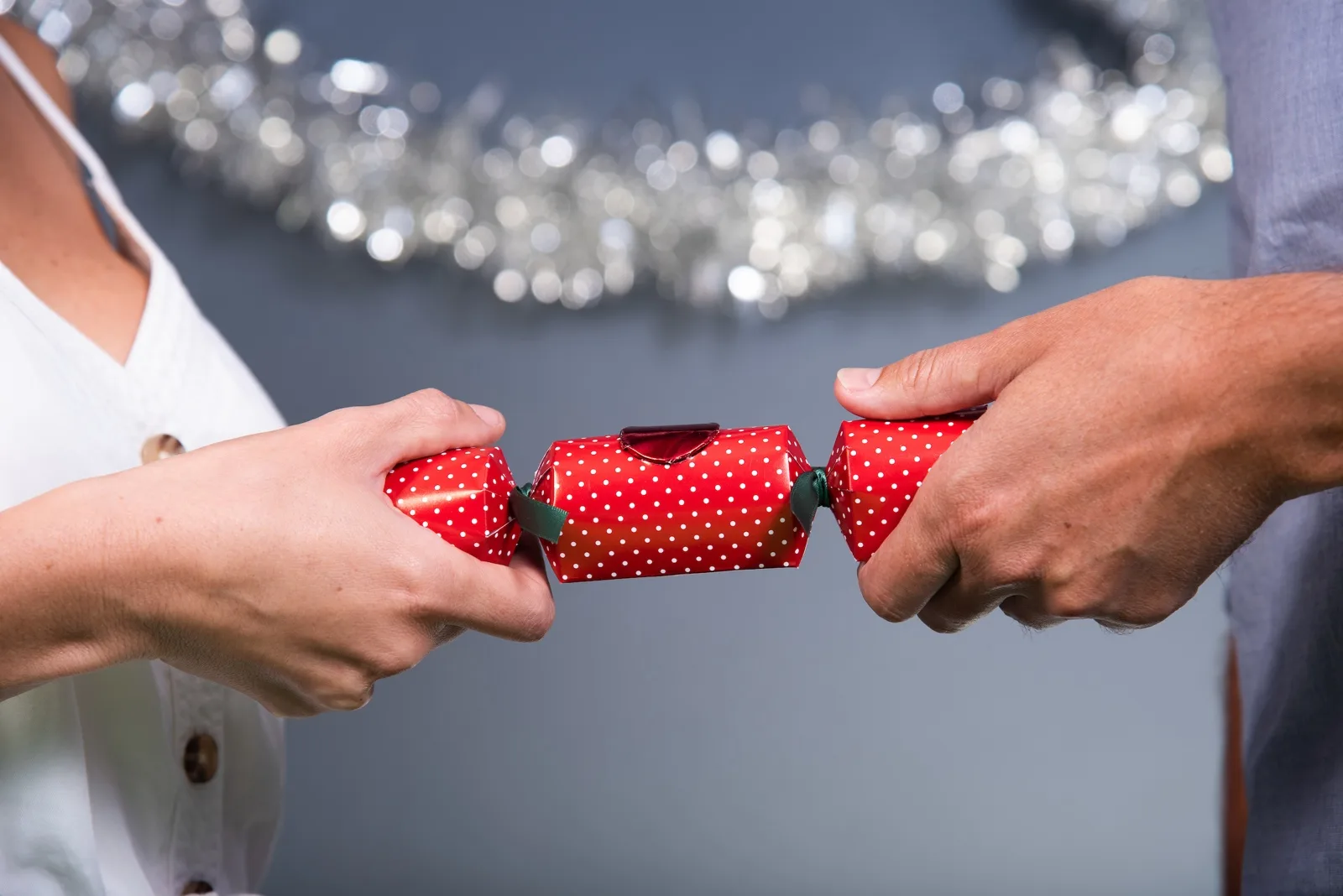 Two people pulling a red polka dot Crackertoa Christmas cracker in front of silver tinsel