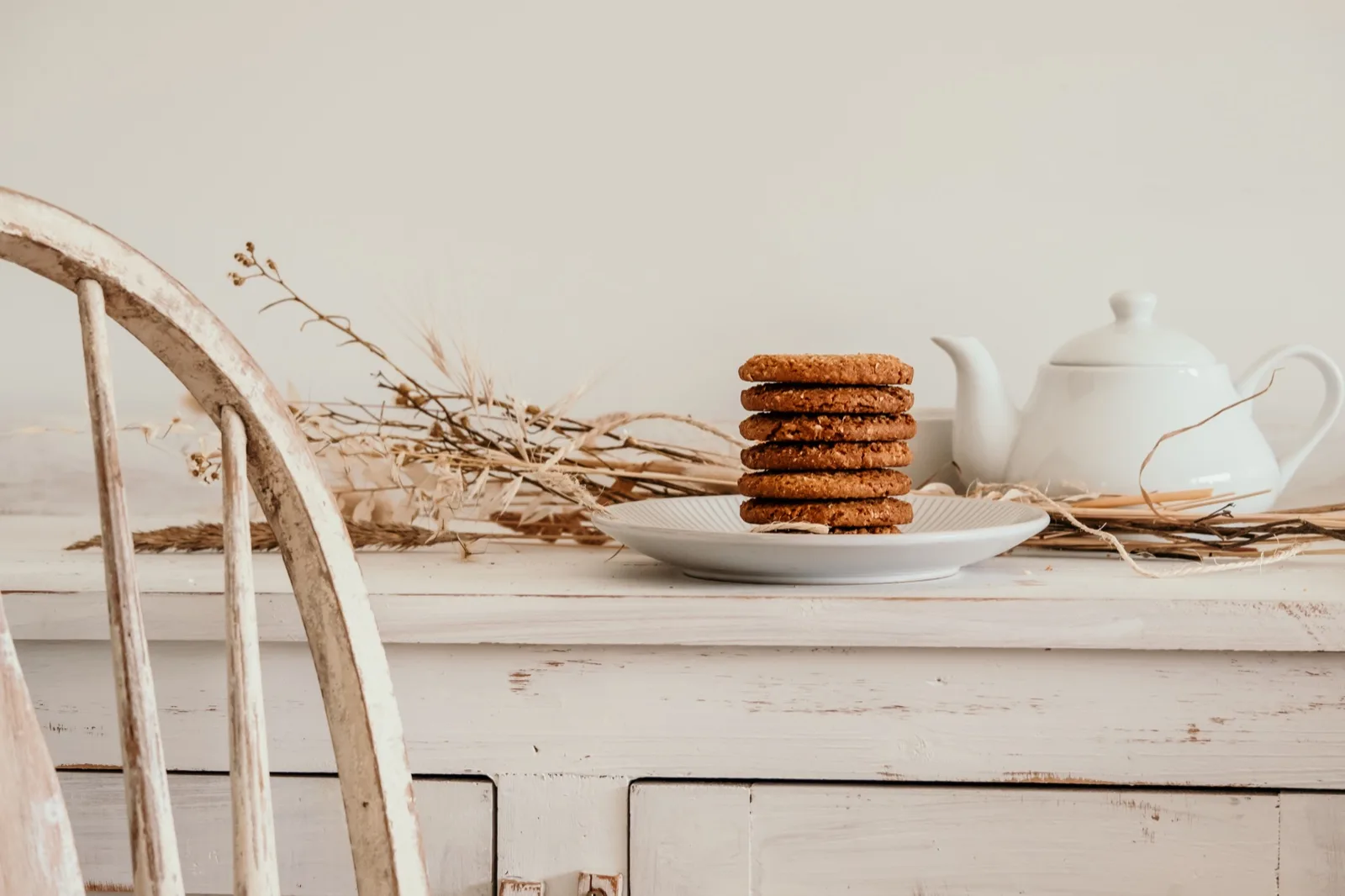 Stack of six hard round golden oat biscuits on a plate beside a white teapot on a whitewas