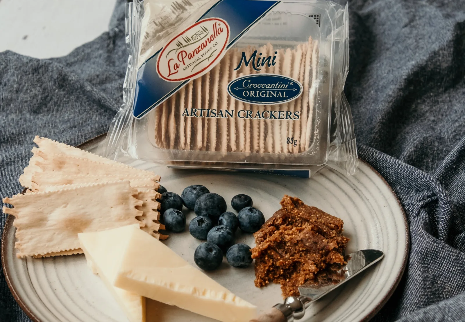 Slab of dark fig paste with a cheese knife beside a wedge of hard cheese, blueberries and artisan crackers