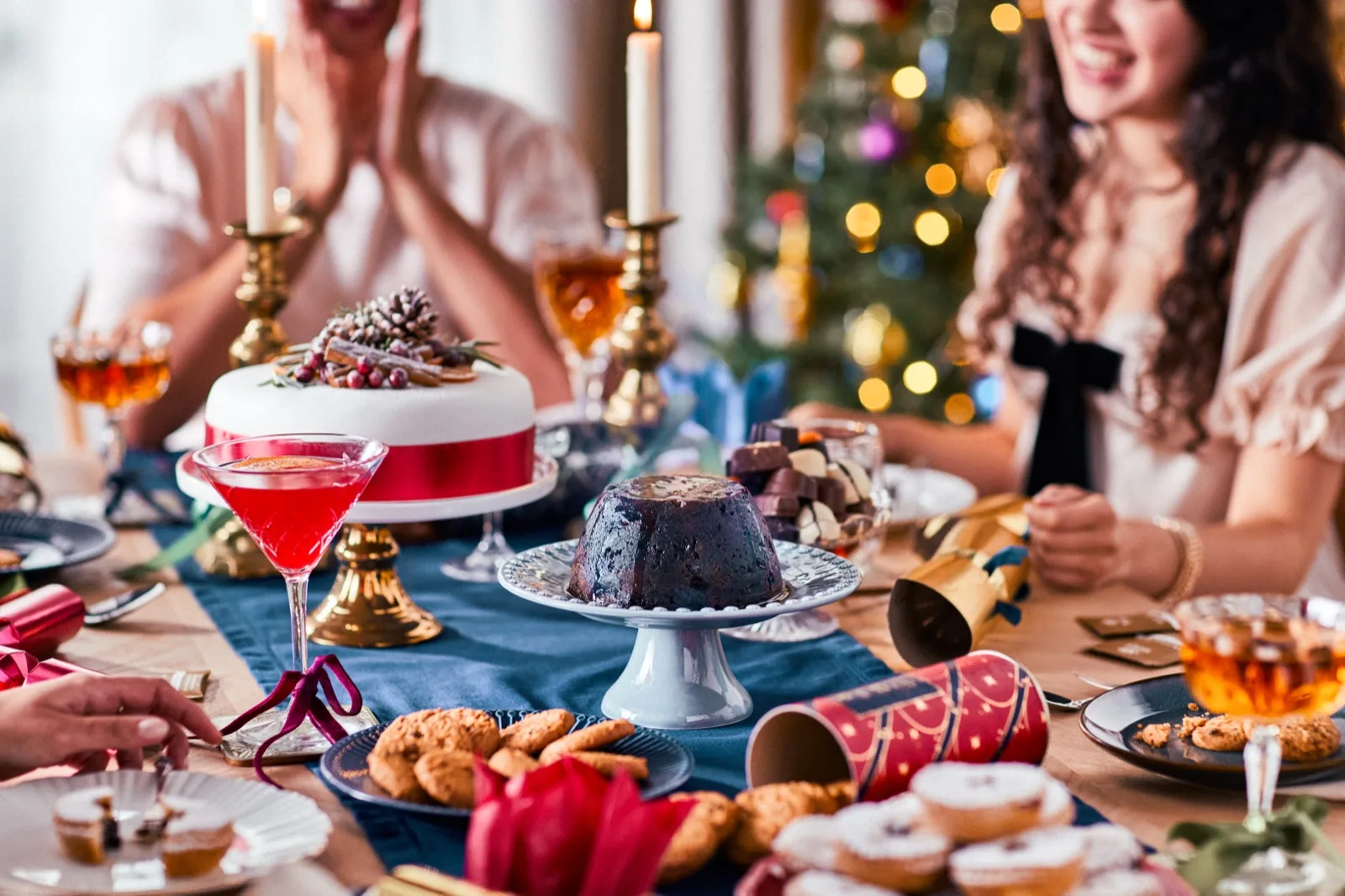 A pink Christmas drink in a coupe on a festive table set with Christmas cake, pudding and 