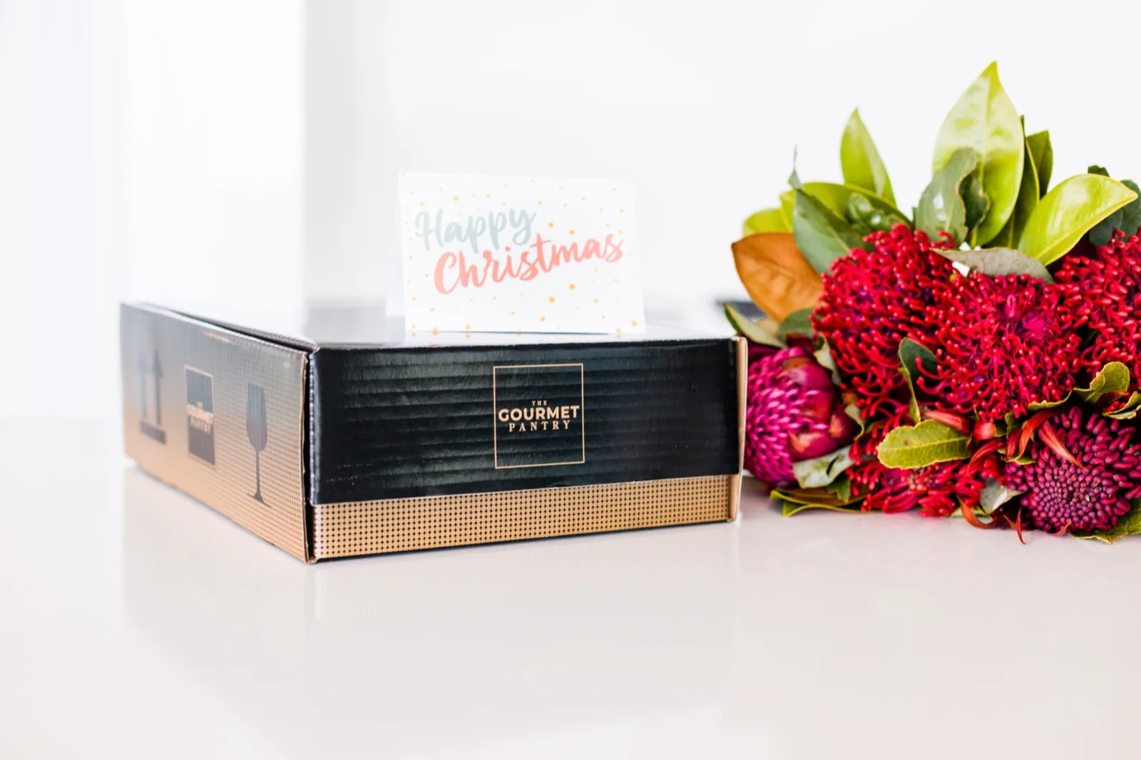 A sealed black and gold Gourmet Pantry hamper carton with a printed Happy Christmas gift card standing on top