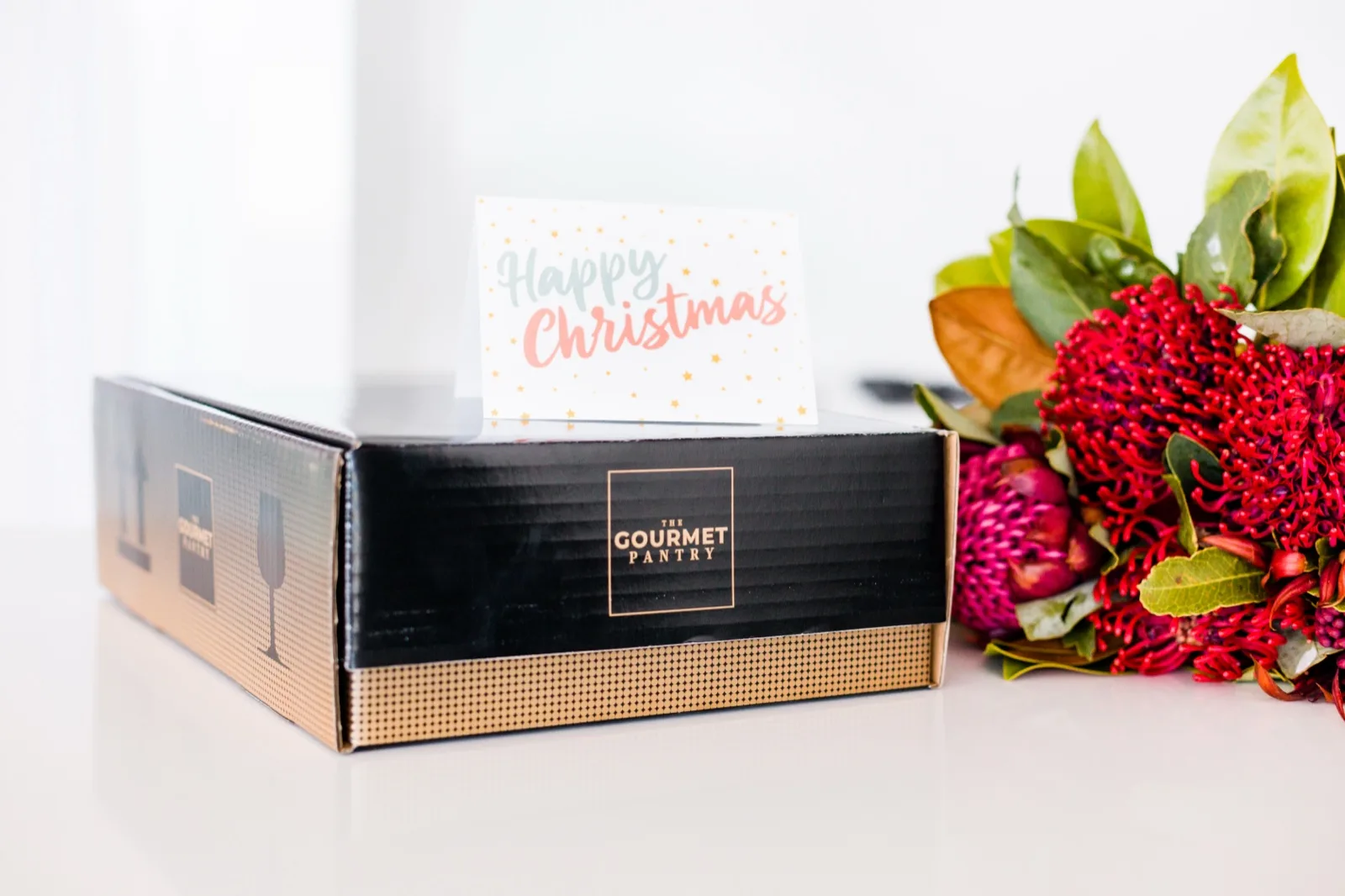A packed The Gourmet Pantry shipping carton with a Happy Christmas card standing on top and waratahs beside