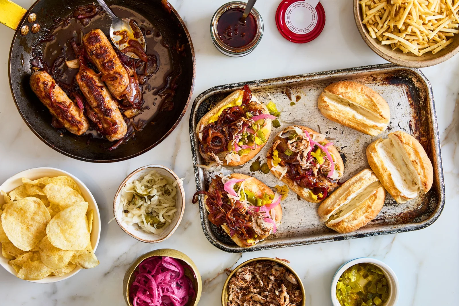 Ottolenghi glazed pork sausages in a pan with caramelised red onion, rolls, crisps and pic