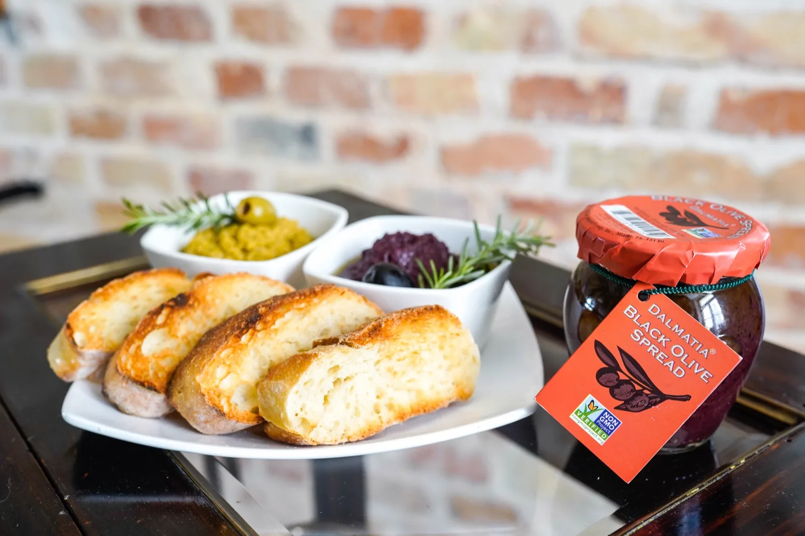 Bowls of black and green olive spread with toasted baguette beside a Dalmatia black olive 