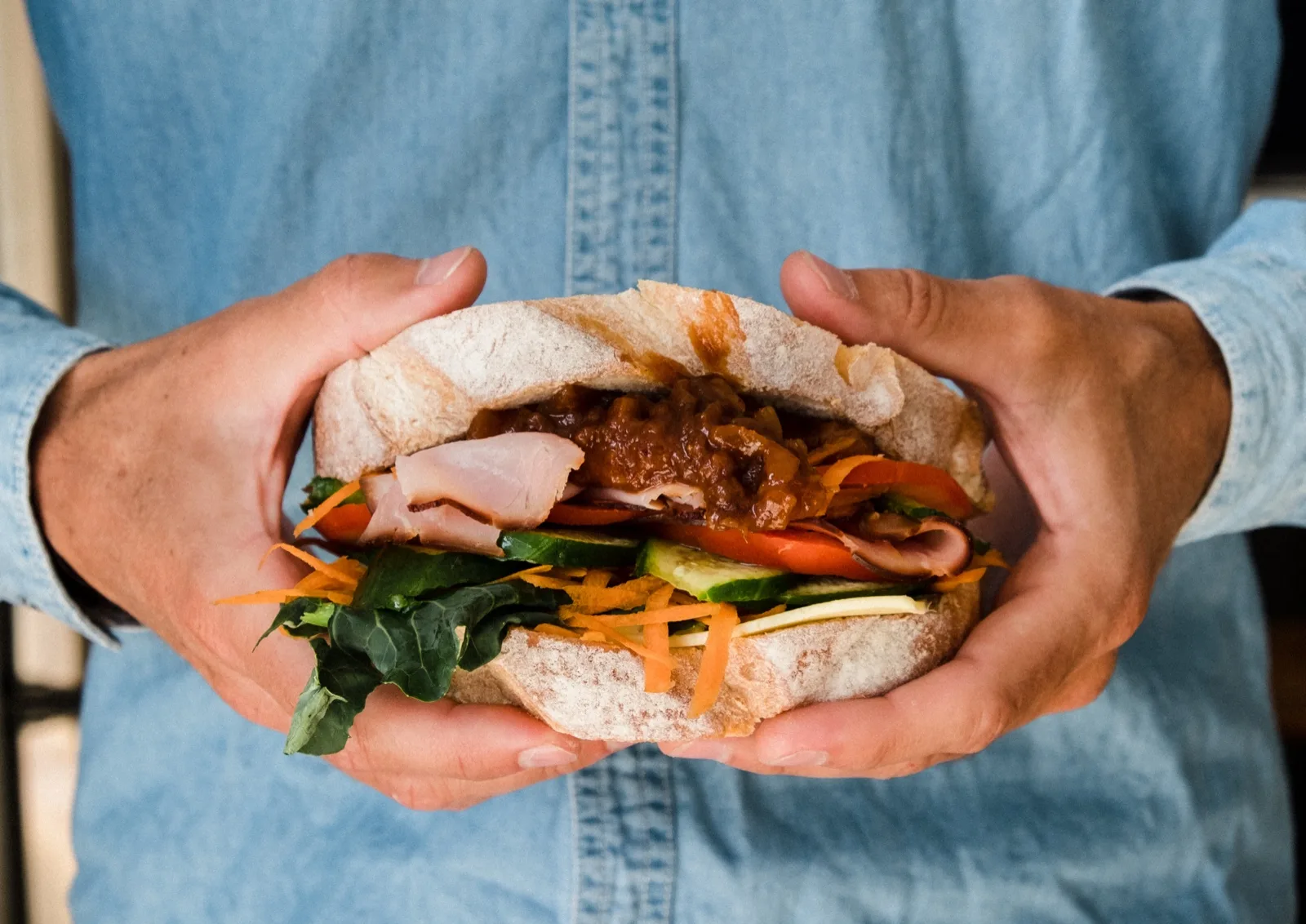 Hands holding a crusty roll of ham, cheese, tomato, cucumber and carrot topped with a spoo